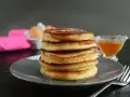 Pila di pancake senza latte, dorati e soffici, con sciroppo d'acero e un tovagliolo rosa sullo sfondo.