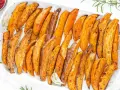 Patate dolci al forno, tagliate a spicchi e condite con erbe aromatiche, servite su un piatto bianco.