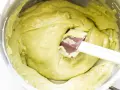 A spatula mixes a smooth, pale green pistachio cream in a stainless steel bowl.