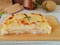 Una fetta di parmigiana di patate dorata e filante, con strati di patate, prosciutto e besciamella, guarnita con rosmarino.