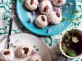 Biscotti al cocco spolverati di zucchero a velo, decorati con mandorle e fiori viola, accanto a una tazza di tè alla menta.