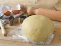 Impasto di pasta frolla con olio, pronto per essere trasformato in biscotti con stampini a forma di stella e fiore. Guscio d'uovo e mattarello completano la scena.