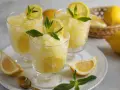 Tre bicchieri di rinfrescante granita al limone, guarniti con fettine di limone e foglie di menta. Limoni freschi in un cestino sullo sfondo.