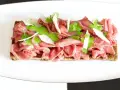 Delicato carpaccio di manzo su crostino integrale, guarnito con crema verde, scaglie di parmigiano e foglioline fresche.