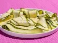 Fettine sottili di zucchine marinate crude, condite con pepe nero, su un piatto bianco, su un tavolo rosa acceso.