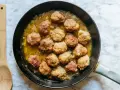 Polpette dorate in padella con cipolle, pronte per essere gustate. Un cucchiaio di legno attende accanto.