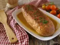 Polpettone dorato in padella, guarnito con erba cipollina, su un piatto bianco con salsa, accanto a pomodorini e una spatola di legno.