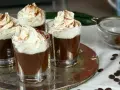 Quattro bicchierini di budino al cioccolato, guarniti con panna montata e cacao in polvere, su un vassoio d'argento.