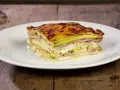 Una fetta di parmigiana di zucchine bianca, stratificata con prosciutto e formaggio filante, su un piatto bianco.