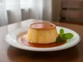 Budino fatto in casa con caramello dorato e una fogliolina di menta fresca, servito su un piatto bianco.