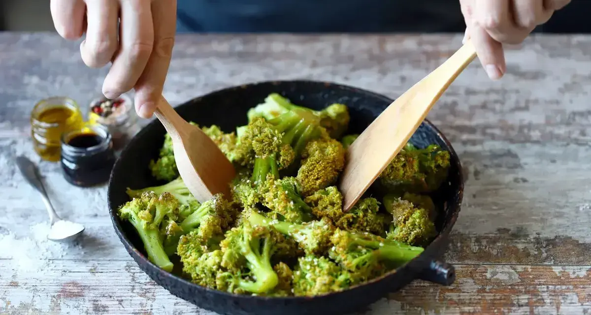 Broccoli in padella, condito con olio e spezie, viene mescolato con due palette di legno.