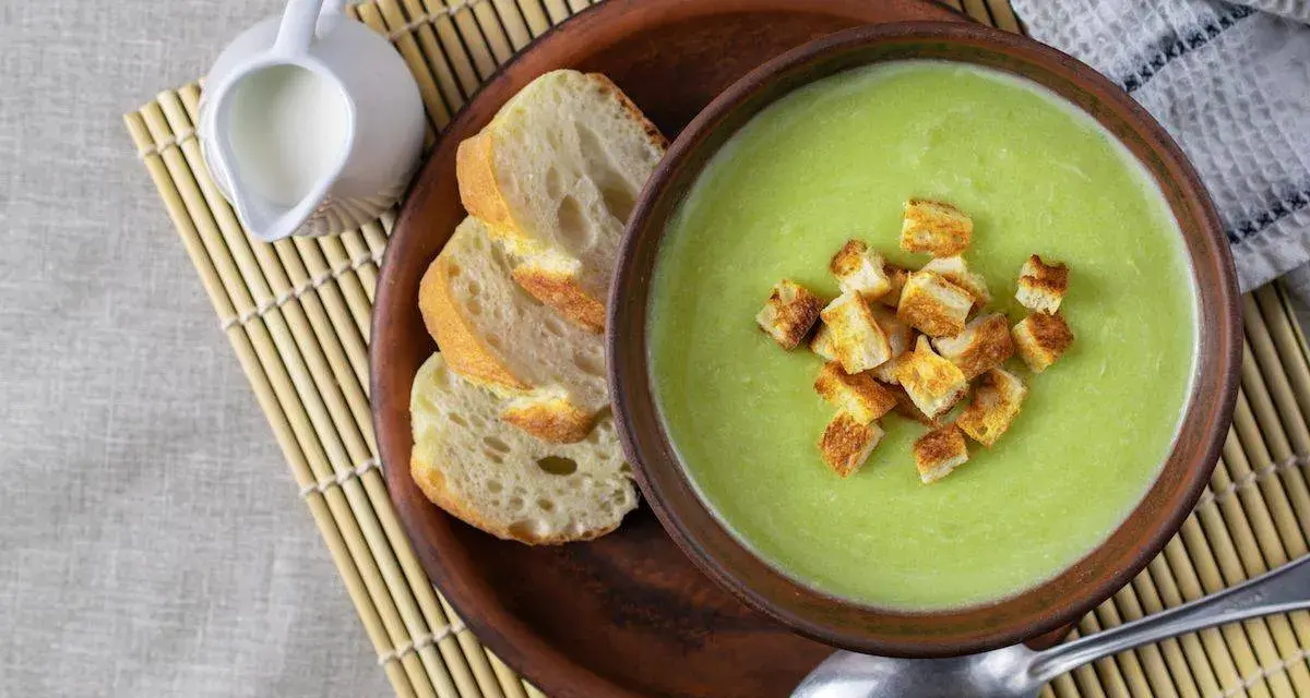 Una ciotola di cremosa crema di zucchine con crostini dorati, accompagnata da pane fresco e una caraffa di latte.