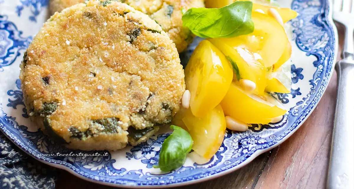 Hamburger di zucchine dorati e croccanti, serviti con pomodorini gialli e basilico fresco su un piatto blu e bianco.