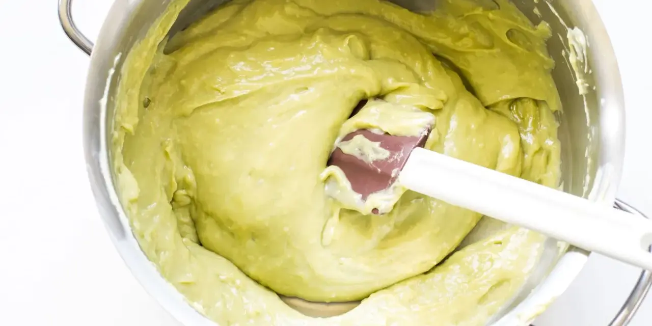 A spatula mixes a smooth, pale green pistachio cream in a stainless steel bowl.