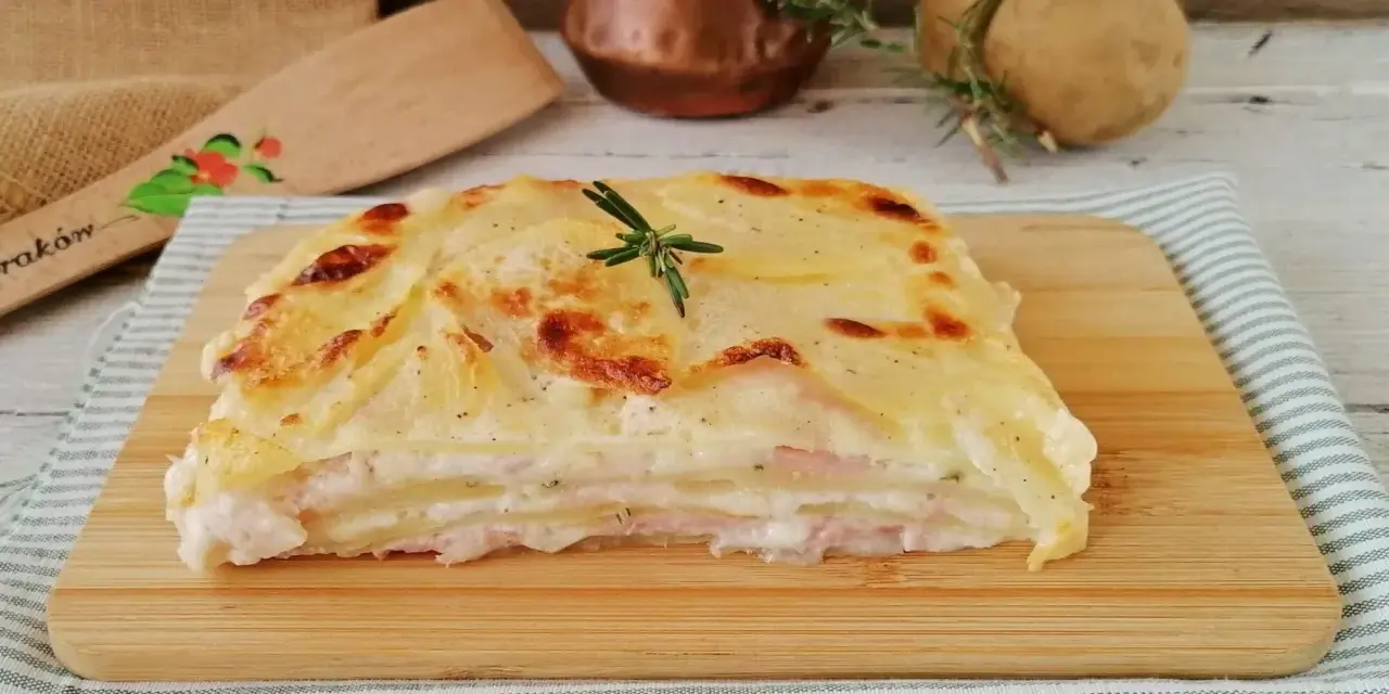 Una fetta di parmigiana di patate dorata e filante, con strati di patate, prosciutto e besciamella, guarnita con rosmarino.