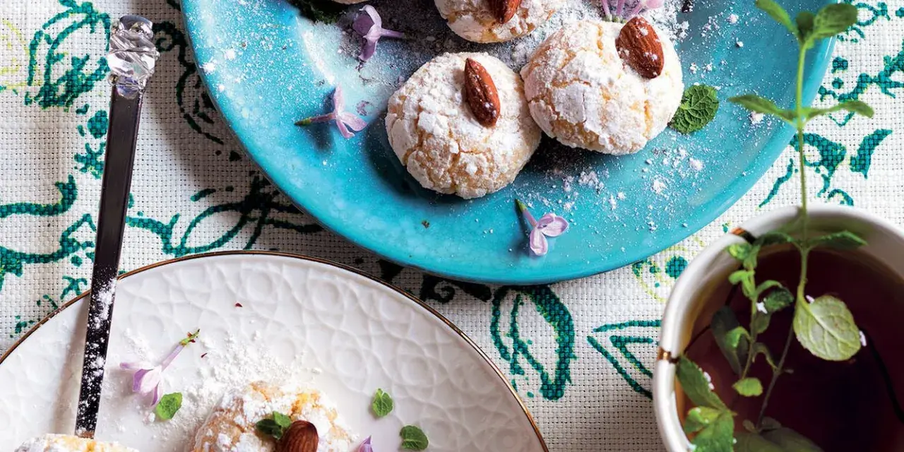 Biscotti al cocco spolverati di zucchero a velo, decorati con mandorle e fiori viola, accanto a una tazza di tè alla menta.