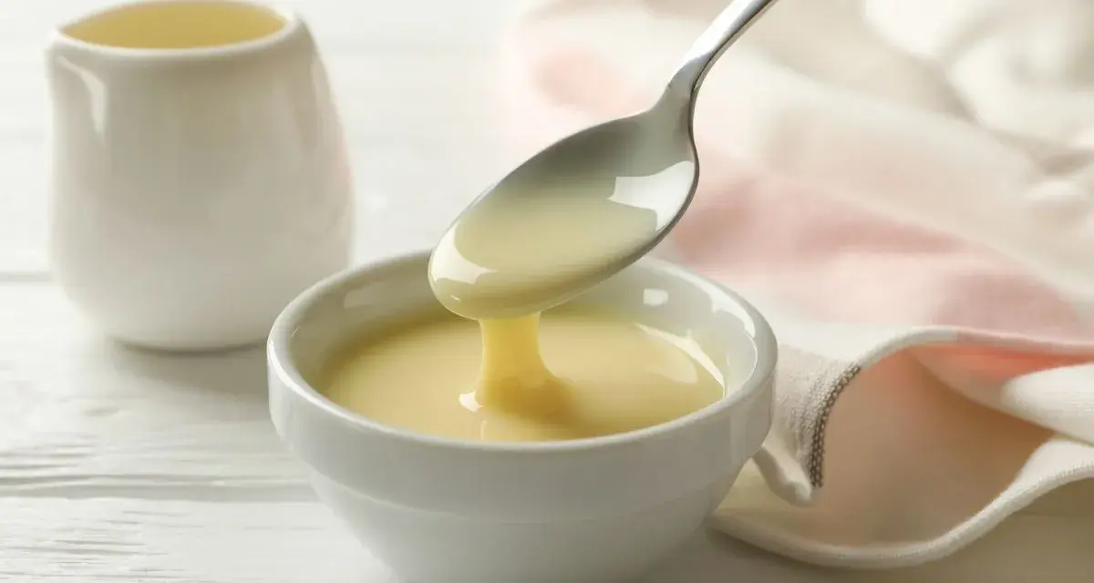 Latte condensato denso e cremoso, perfetto per dolci fatti col Bimby, cola da un cucchiaio in una ciotola bianca.