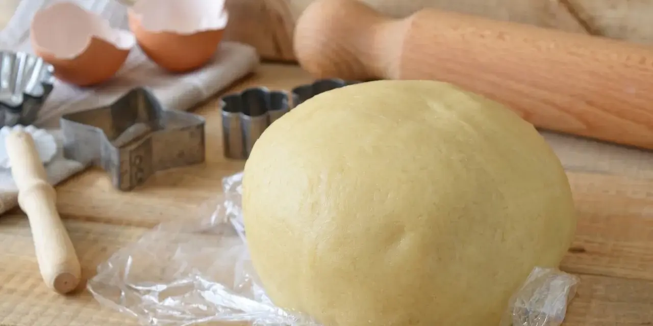 Impasto di pasta frolla con olio, pronto per essere trasformato in biscotti con stampini a forma di stella e fiore. Guscio d'uovo e mattarello completano la scena.