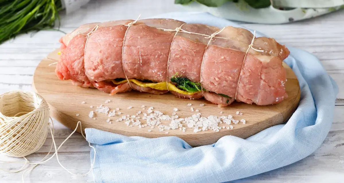 Un arrosto al forno pronto per essere cucinato, legato con spago, su un tagliere con sale grosso e verdure.