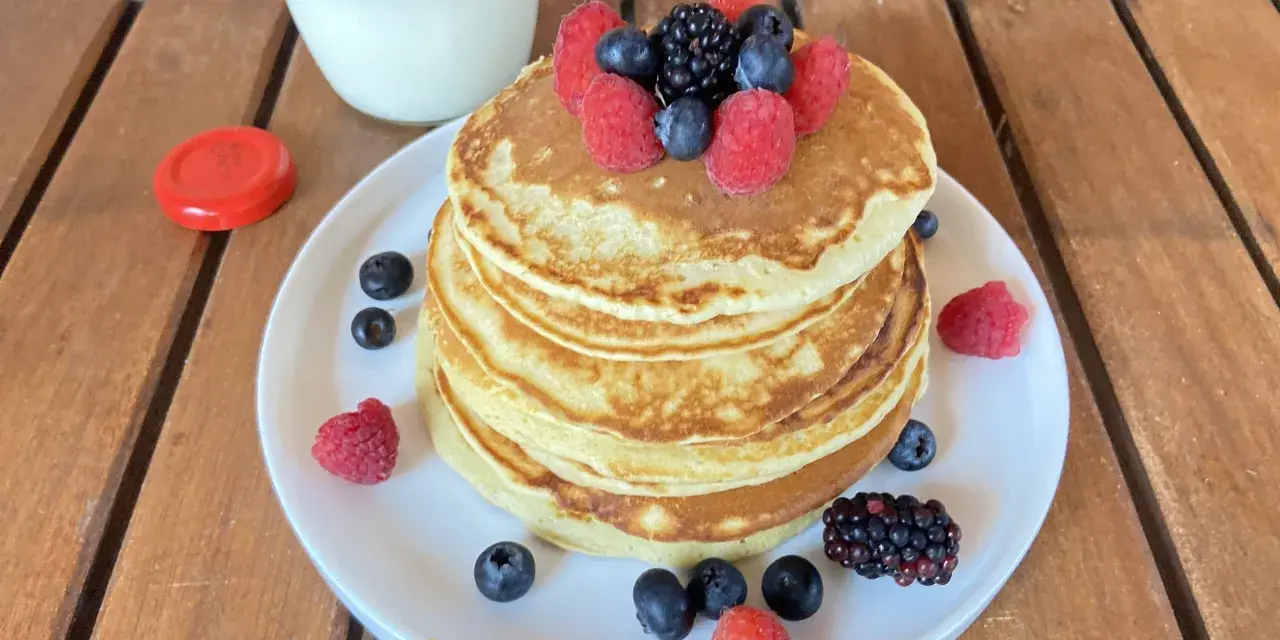 Pila di pancake senza zucchero guarnita con frutti di bosco freschi, accompagnata da un bicchiere di latte.
