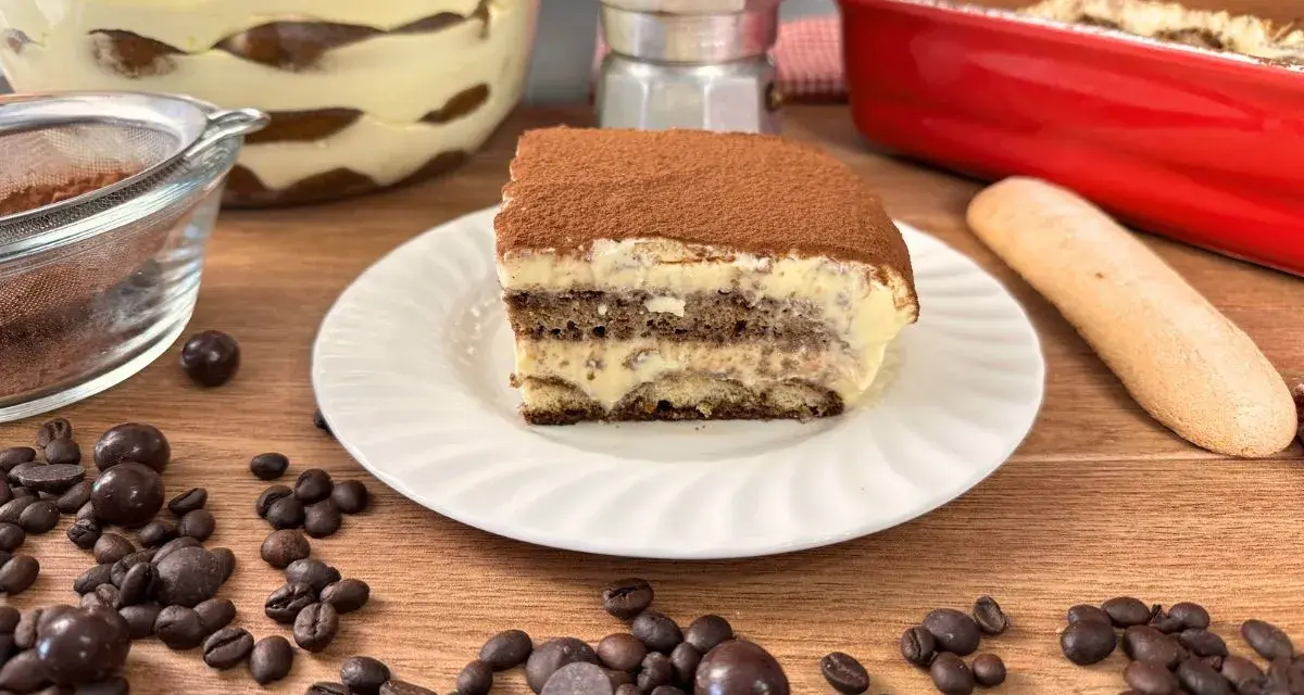 Una fetta di tiramisù con uova pastorizzate, cosparso di cacao, su un piatto bianco, circondato da chicchi di caffè e cioccolatini.