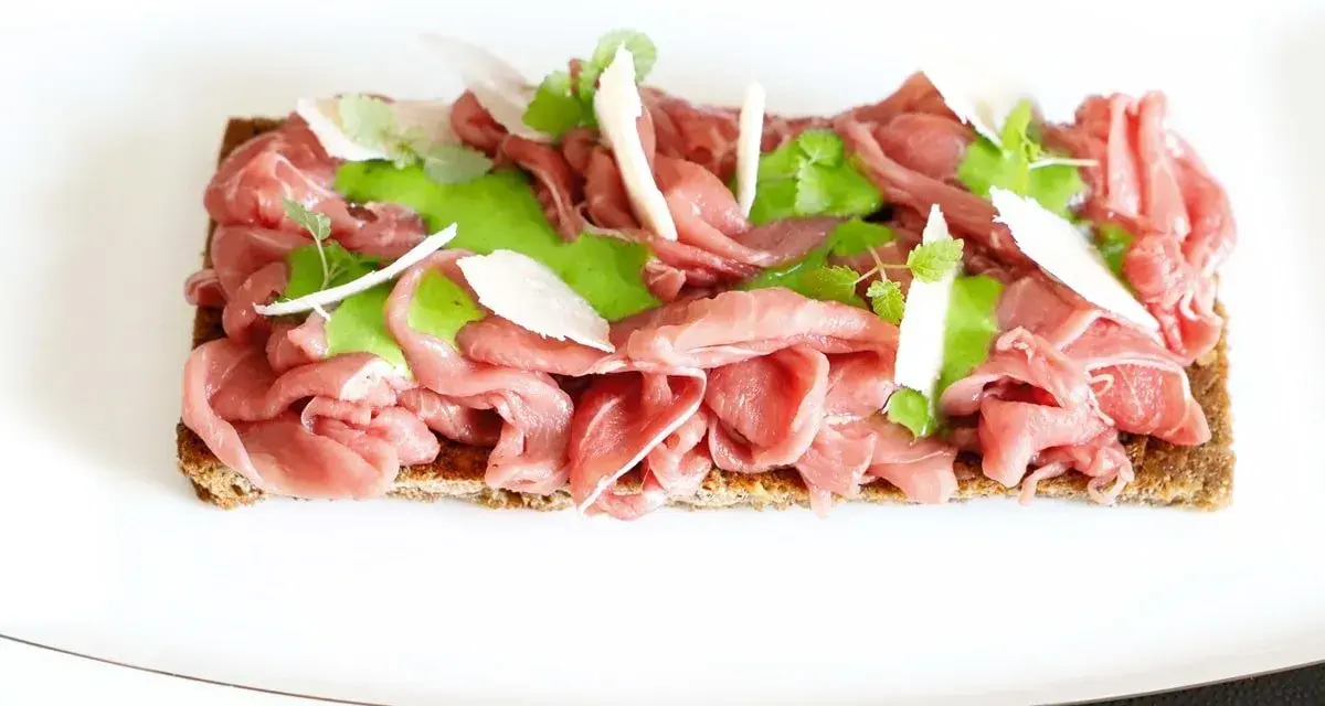Delicato carpaccio di manzo su crostino integrale, guarnito con crema verde, scaglie di parmigiano e foglioline fresche.