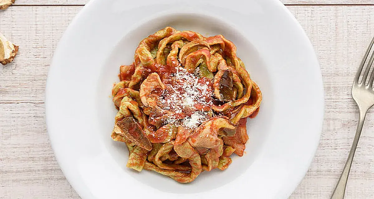 Un piatto di tagliatelle ai funghi, condite con sugo rosso e parmigiano grattugiato, su un tavolo di legno.
