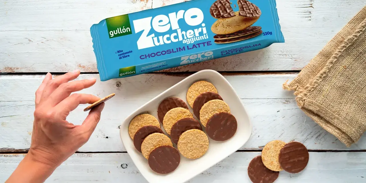 Biscotti senza zucchero Gullón Chocoslim Latte, alcuni ricoperti di cioccolato, su un piatto bianco. Una mano ne prende uno.