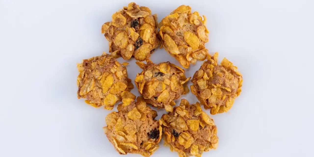Biscotti croccanti, simili a rose del deserto, con fiocchi di mais e uvetta, disposti a cerchio su sfondo bianco.