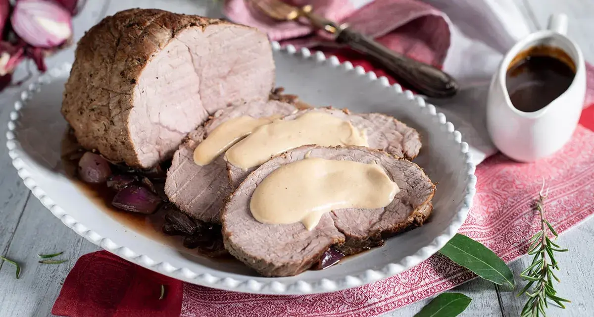 Arrosto di vitello morbido con sughetto benedetta, servito su un piatto bianco con cipolle rosse e rosmarino.