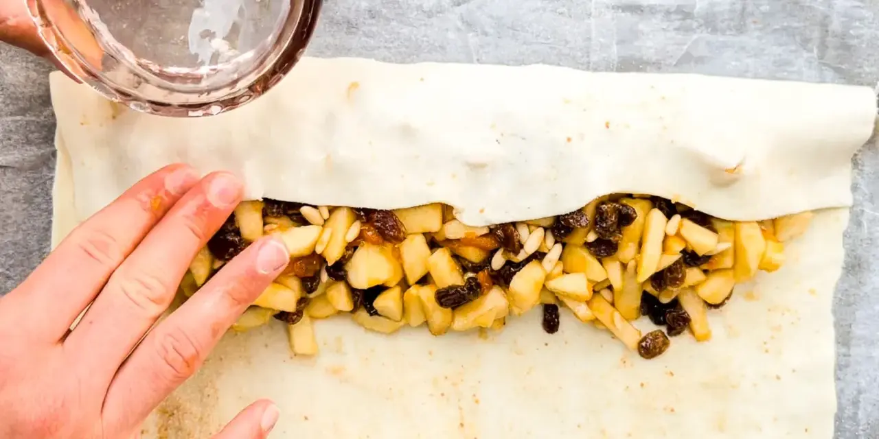 Mani che preparano uno strudel di mele con pasta sfoglia, riempiendolo con mele a cubetti, uvetta e pinoli.
