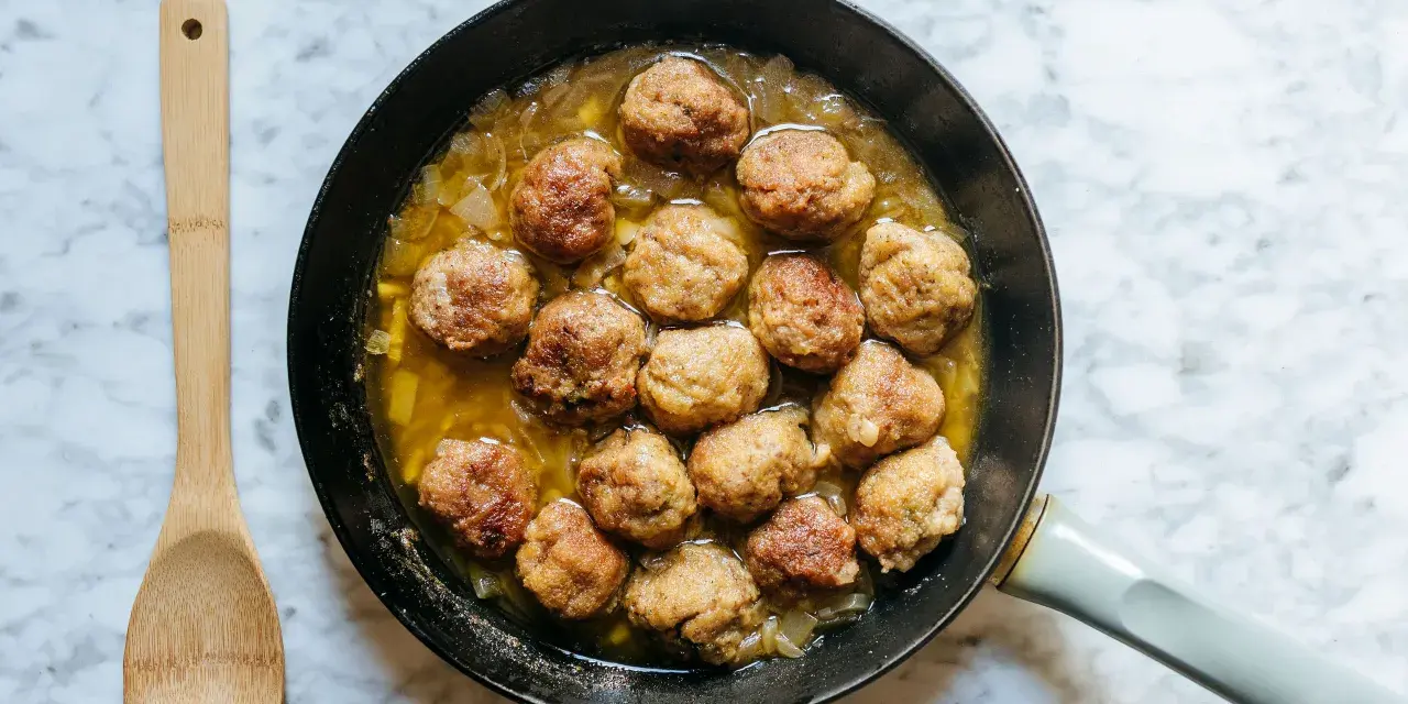 Polpette dorate in padella con cipolle, pronte per essere gustate. Un cucchiaio di legno attende accanto.