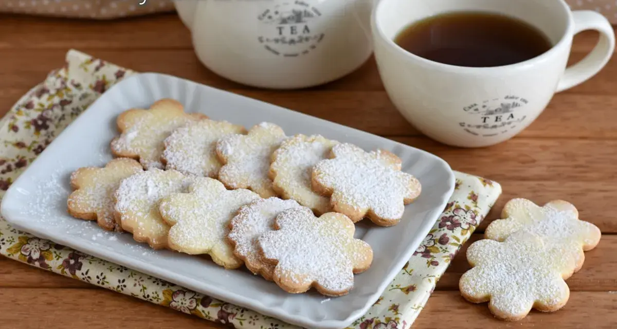 Biscotti con farina di riso a forma di fiore, spolverati di zucchero a velo, accanto a una tazza di tè e una teiera.