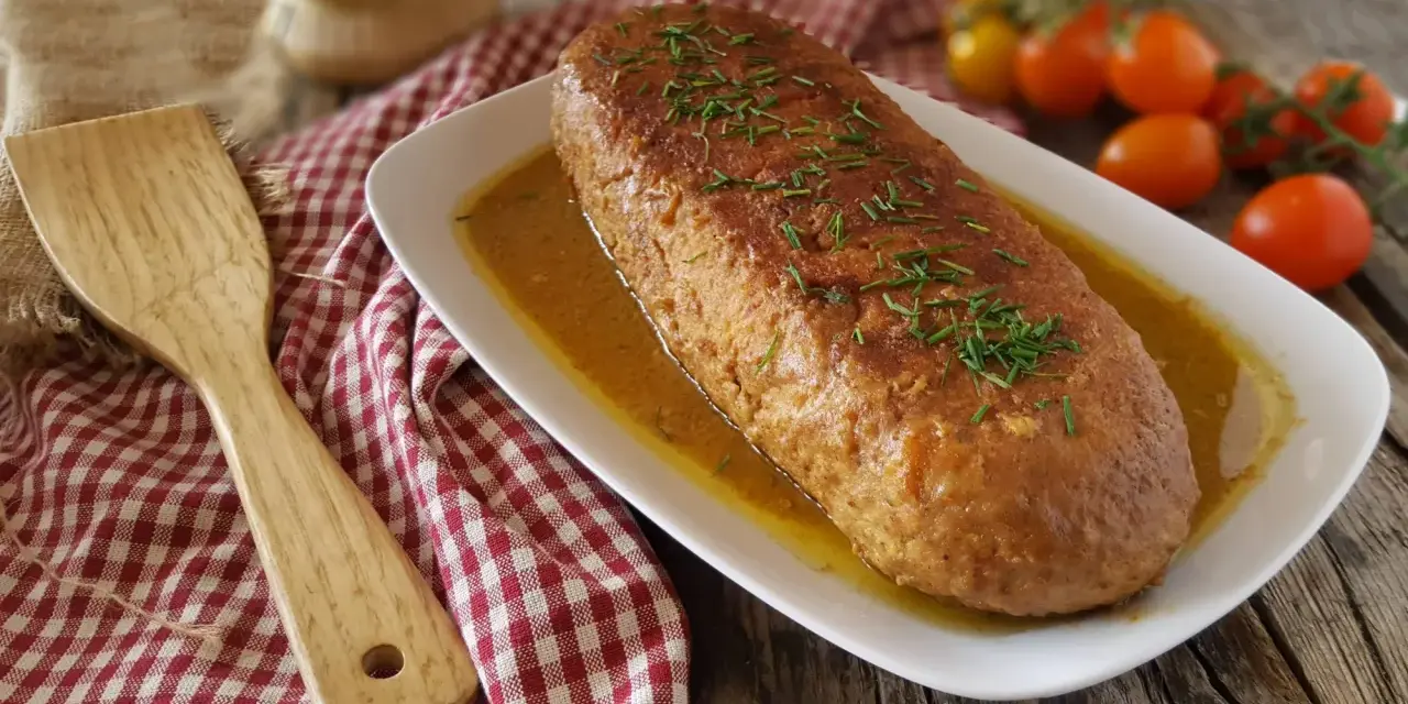 Polpettone dorato in padella, guarnito con erba cipollina, su un piatto bianco con salsa, accanto a pomodorini e una spatola di legno.