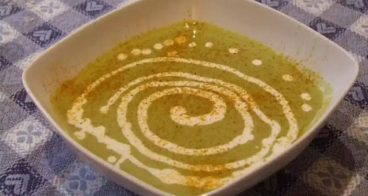 Una ciotola quadrata bianca contiene una vellutata crema di piselli, decorata con un vortice di panna e una spolverata di spezie.