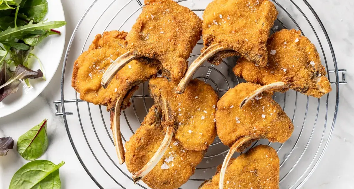 Deliziose costolette d'agnello fritte, dorate e croccanti, cosparse di sale grosso, su una griglia. Accanto, un'insalata fresca.