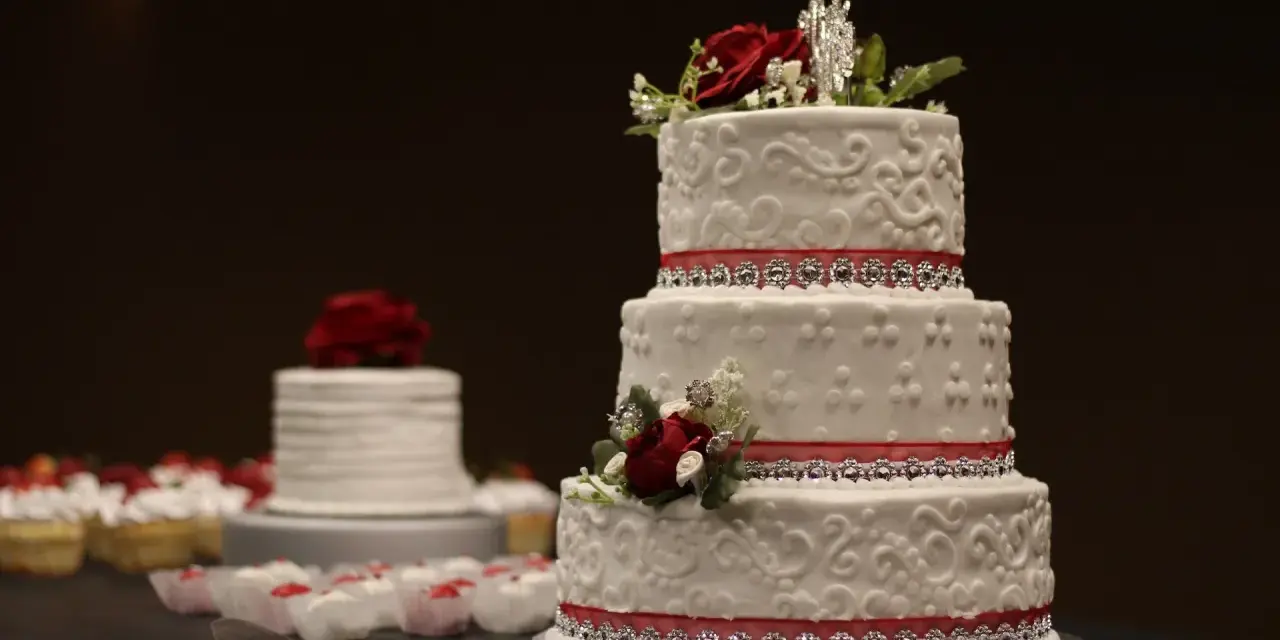 Torta nuziale a tre piani decorata con pasta di zucchero, rose rosse e nastri scintillanti. Cupcake e un'altra torta sullo sfondo.