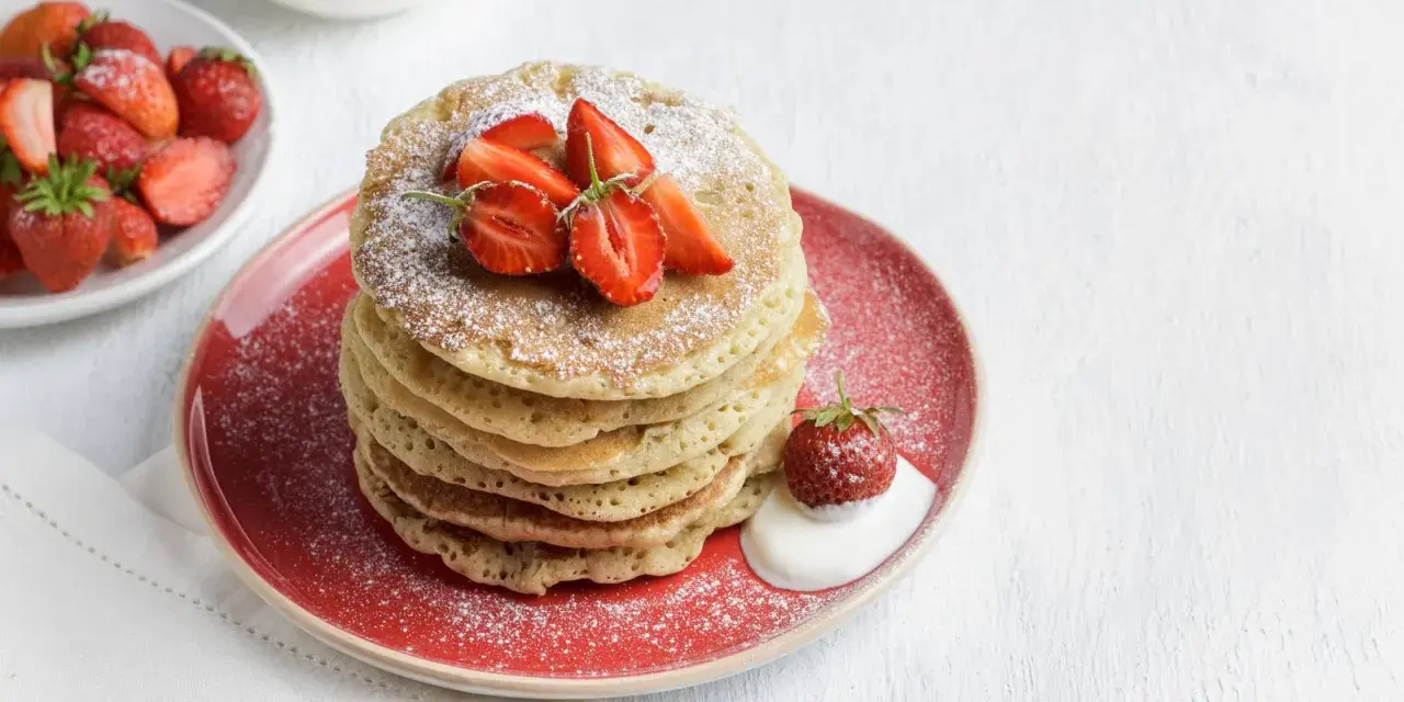 Pila di pancake senza glutine guarniti con fragole fresche e zucchero a velo, serviti su un piatto rosso con un cucchiaio di panna.