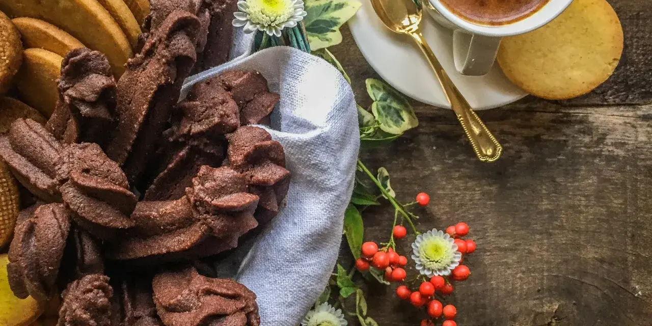 Cesto pieno di biscotti fatti in casa al cioccolato e alla vaniglia, accanto a una tazzina di caffè e bacche rosse.