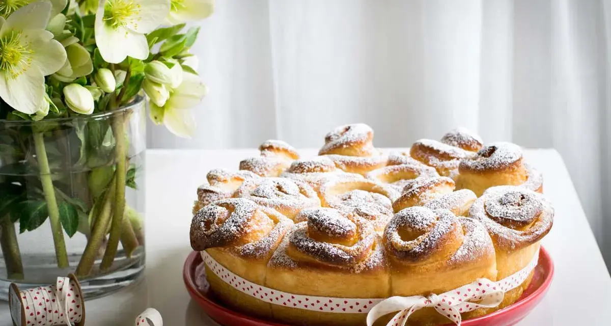 Una deliziosa torta di rose, cosparsa di zucchero a velo, su un piatto rosso, con un nastro a cuoricini. Accanto, un vaso con fiori bianchi.