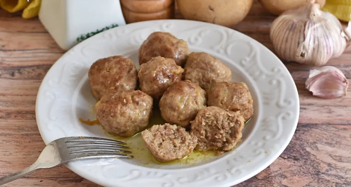 Piatto di polpette in bianco, morbide e succulente, pronte per essere gustate. Sullo sfondo patate e aglio.