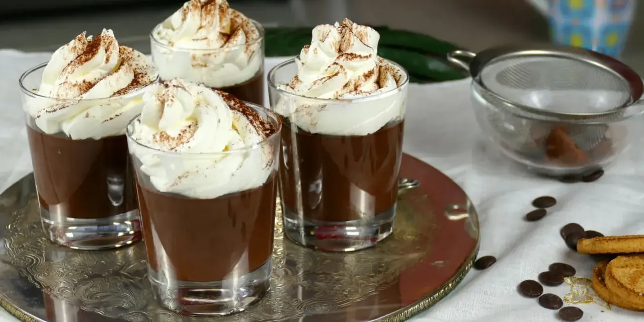 Quattro bicchierini di budino al cioccolato, guarniti con panna montata e cacao in polvere, su un vassoio d'argento.