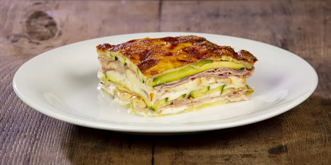 Una fetta di parmigiana di zucchine bianca, stratificata con prosciutto e formaggio filante, su un piatto bianco.