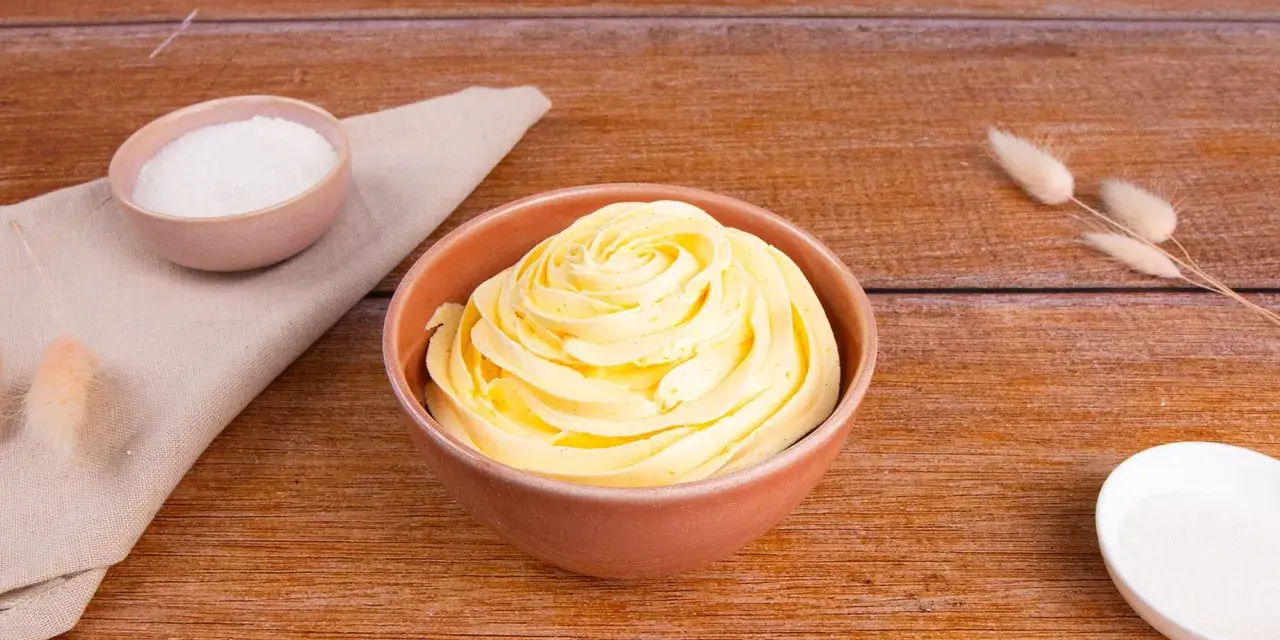 Una ciotola piena di soffice crema al burro, pronta per decorare torte. Accanto, ciotoline con zucchero e decorazioni naturali.
