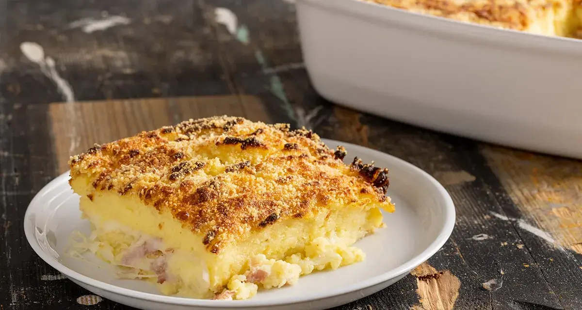 Una fetta di sformato di patate dorato e croccante, con prosciutto e formaggio filante, servita su un piatto bianco.