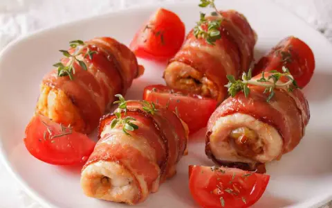 Involtini di pollo avvolti in pancetta croccante, guarniti con timo fresco e serviti con spicchi di pomodoro.