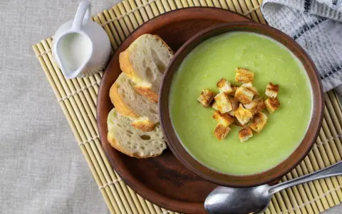 Una ciotola di cremosa crema di zucchine con crostini dorati, accompagnata da pane fresco e una caraffa di latte.
