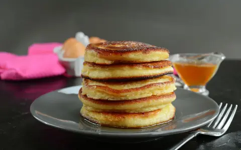 Pila di pancake senza latte, dorati e soffici, con sciroppo d'acero e un tovagliolo rosa sullo sfondo.