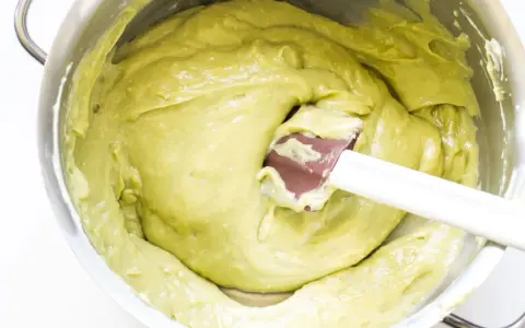 A spatula mixes a smooth, pale green pistachio cream in a stainless steel bowl.