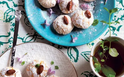 Biscotti al cocco spolverati di zucchero a velo, decorati con mandorle e fiori viola, accanto a una tazza di tè alla menta.